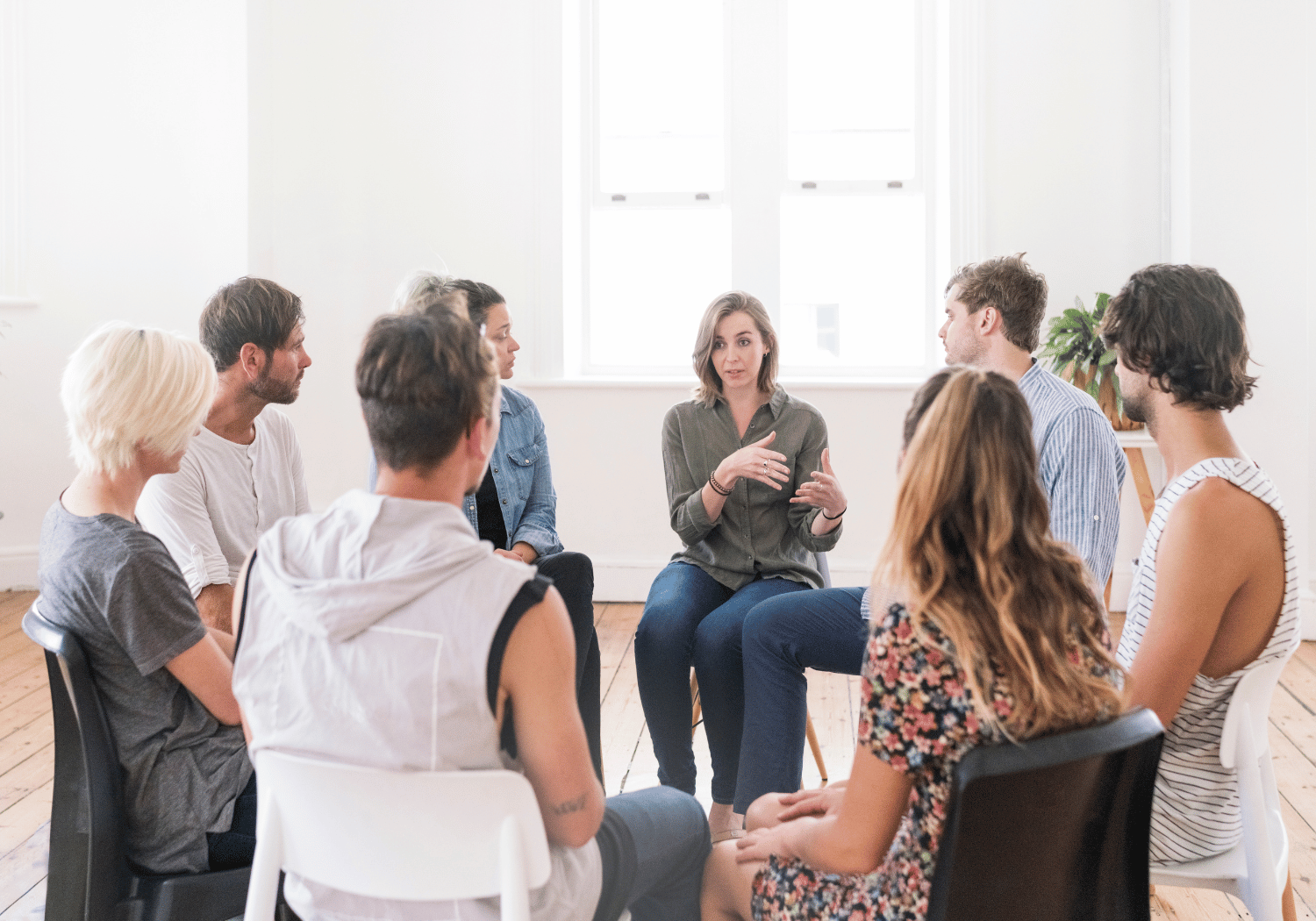 Group of adults sitting in a circle in a bright room, a woman in a green shirt speaks and gestures in the center.